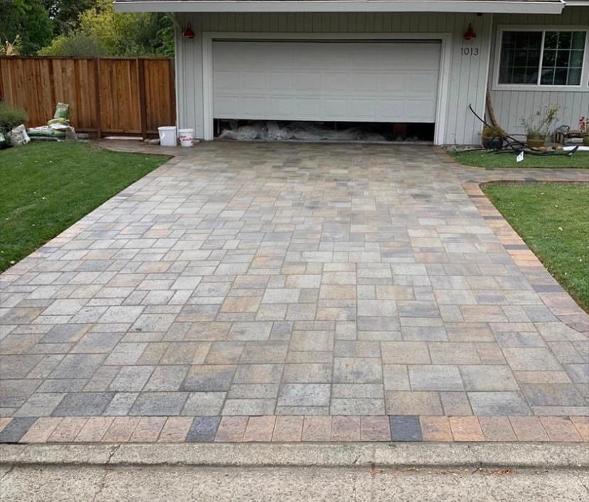 Driveway-Pavers-Service-2 - Three Rocks Pavers, Inc. A driveway with a pattern of gray and beige paving stones leads to a white garage door. A wooden fence and grass are on the sides.