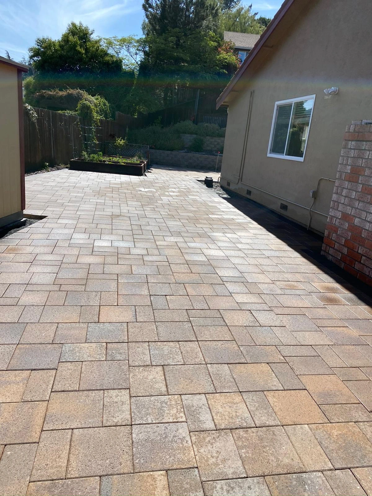 Driveway-Pavers-Service-5 - Three Rocks Pavers, Inc. A sunlit backyard with a spacious stone-paved patio, raised garden bed, and tan brick house on the right.