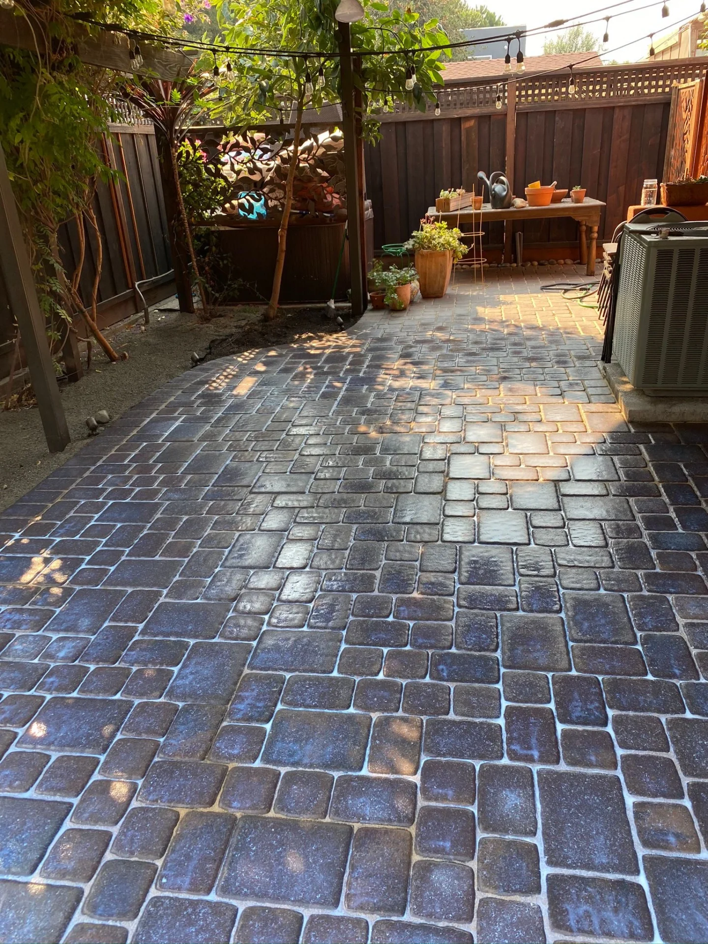 Patio-Pavers-Service-11 - Three Rocks Pavers, Inc. A sunlit backyard patio with decorative cobblestone paving, bordered by plants and a wooden fence, featuring a table with various items and an air conditioning unit on the side.