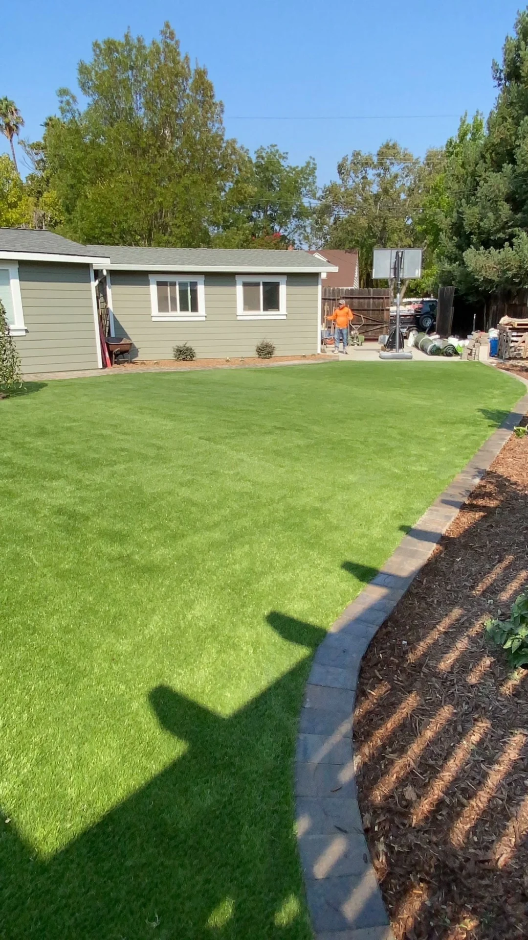 Synthetic-Grass-Service-5 - Three Rocks Pavers, Inc. A backyard with a green lawn, bordered by a stone path. A green house is visible in the background, along with trees and a basketball hoop. Two people are near the hoop.