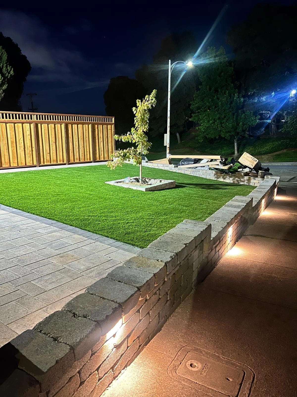 Synthetic-Grass-Service-6 - Three Rocks Pavers, Inc. Night view of a landscaped backyard with illuminated pathways, a small tree centered in a grass area, and a wooden fence in the background.