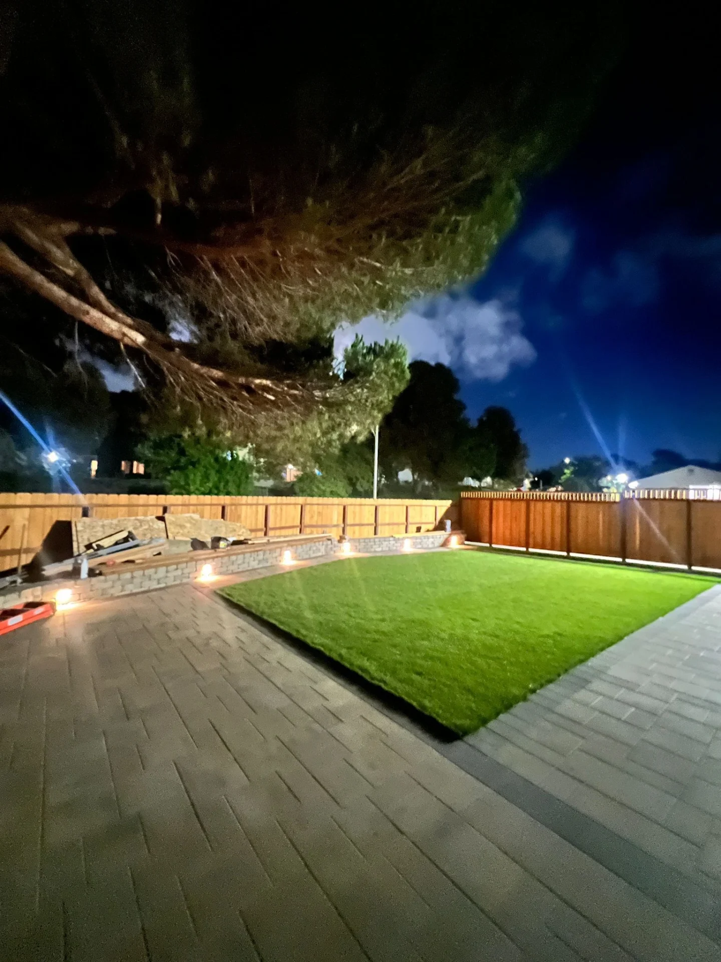 Synthetic-Grass-Service-7 - Three Rocks Pavers, Inc. A well-lit backyard at night with a neatly trimmed lawn, patio pavers, a wooden fence, and trees in the background.