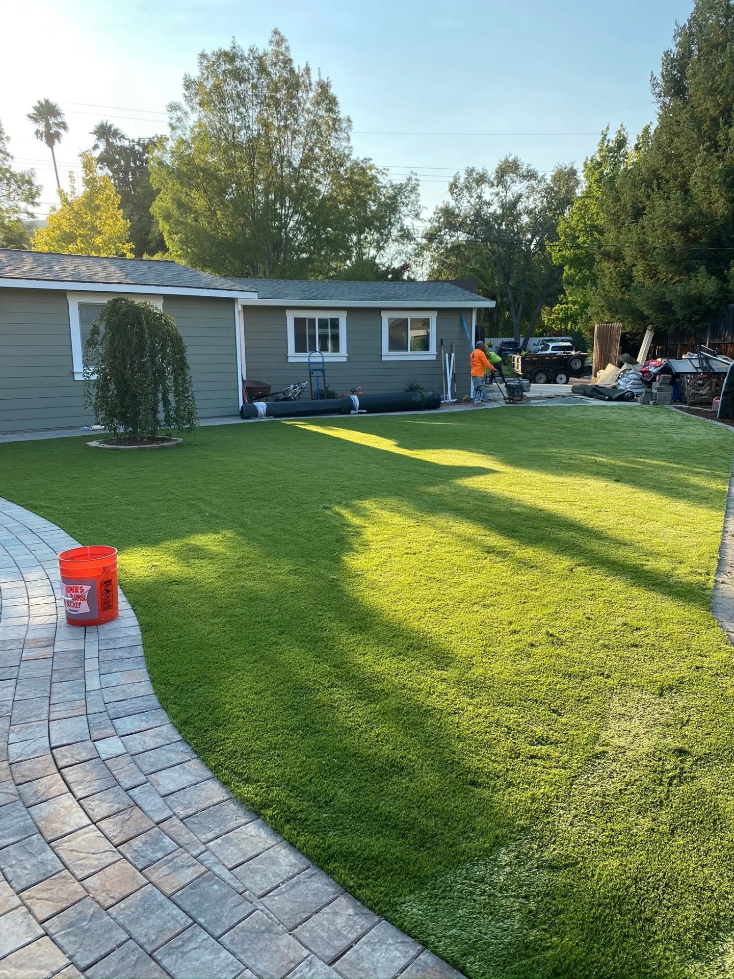 Synthetic-Grass-Service-8 - Three Rocks Pavers, Inc. Backyard with a green lawn, curved stone pathway, gray house, and a person working near the house. A bright orange bucket is on the path. Trees and bushes border the yard.