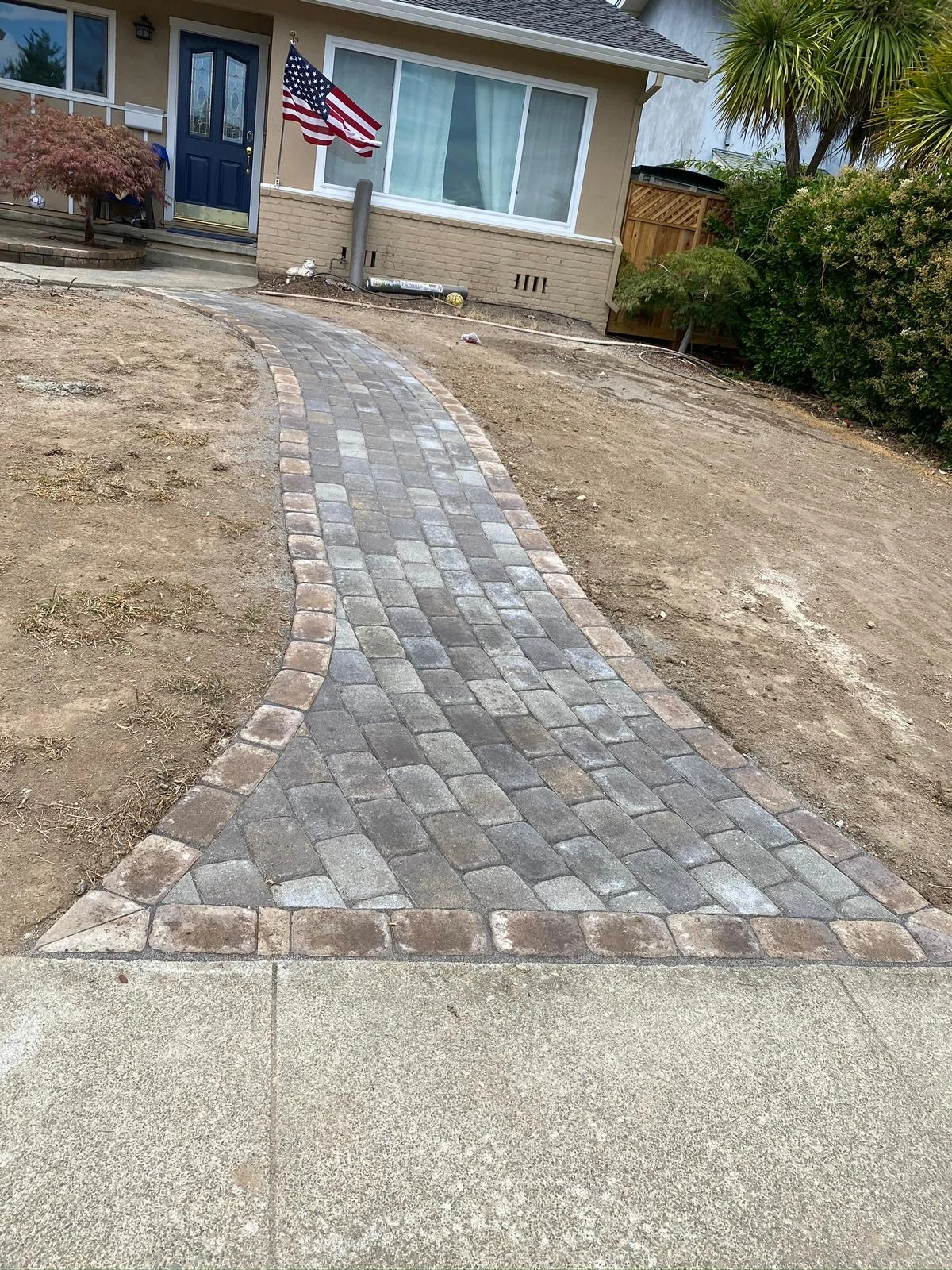 Walkway-Pavers-Service-13 - Three Rocks Pavers, Inc. A newly paved stone walkway leads to a house with a blue door. An American flag is displayed on the front wall. Dry, unlandscaped yard surrounds the path.