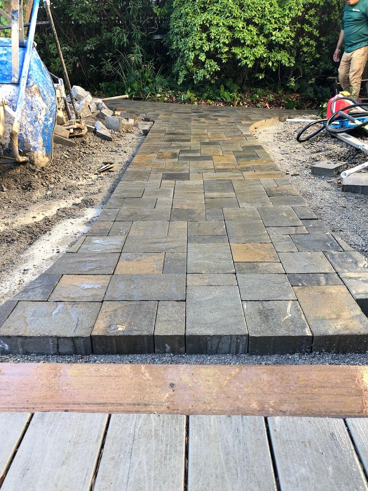 Walkway-Pavers-Service-3 - Three Rocks Pavers, Inc. Brick pathway under construction with gray rectangular pavers. A mixer and tools are visible on the side. Green foliage in the background Outdoor Fire Pits