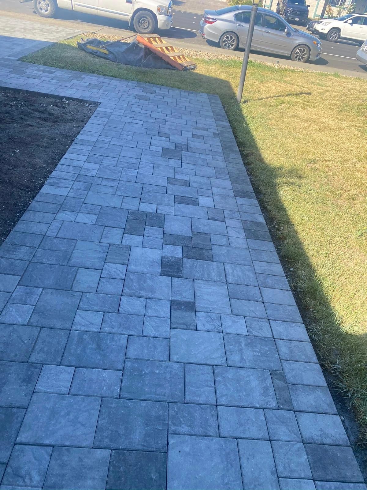 Walkway-Pavers-Service-7 - Three Rocks Pavers, Inc. A paved sidewalk with interlocking gray tiles is bordered by grass. Cars are visible on a nearby street in the background.