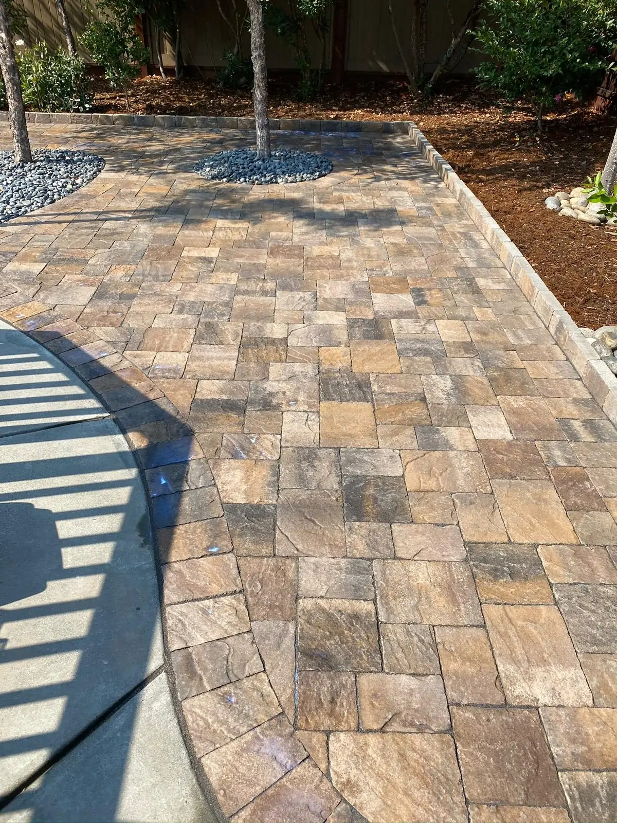 Patio-Pavers-Service-21 1 - Three Rocks Pavers, Inc. Stone patio with interlocking pavers in a mix of earthy tones. Nearby, small trees are planted in circular gravel beds, and there is a concrete border on one side.