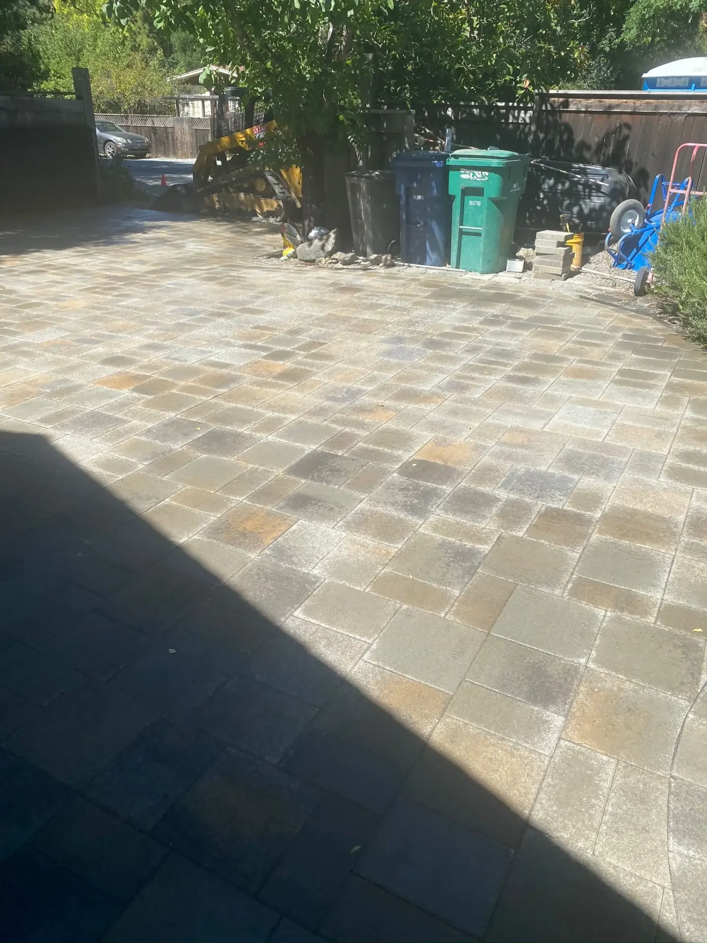 Patio-Pavers-Service-6 1 1 - Three Rocks Pavers, Inc. A sunlit patio with stone pavers. In the background, there are recycling and garbage bins, garden tools, and a shaded area with trees.