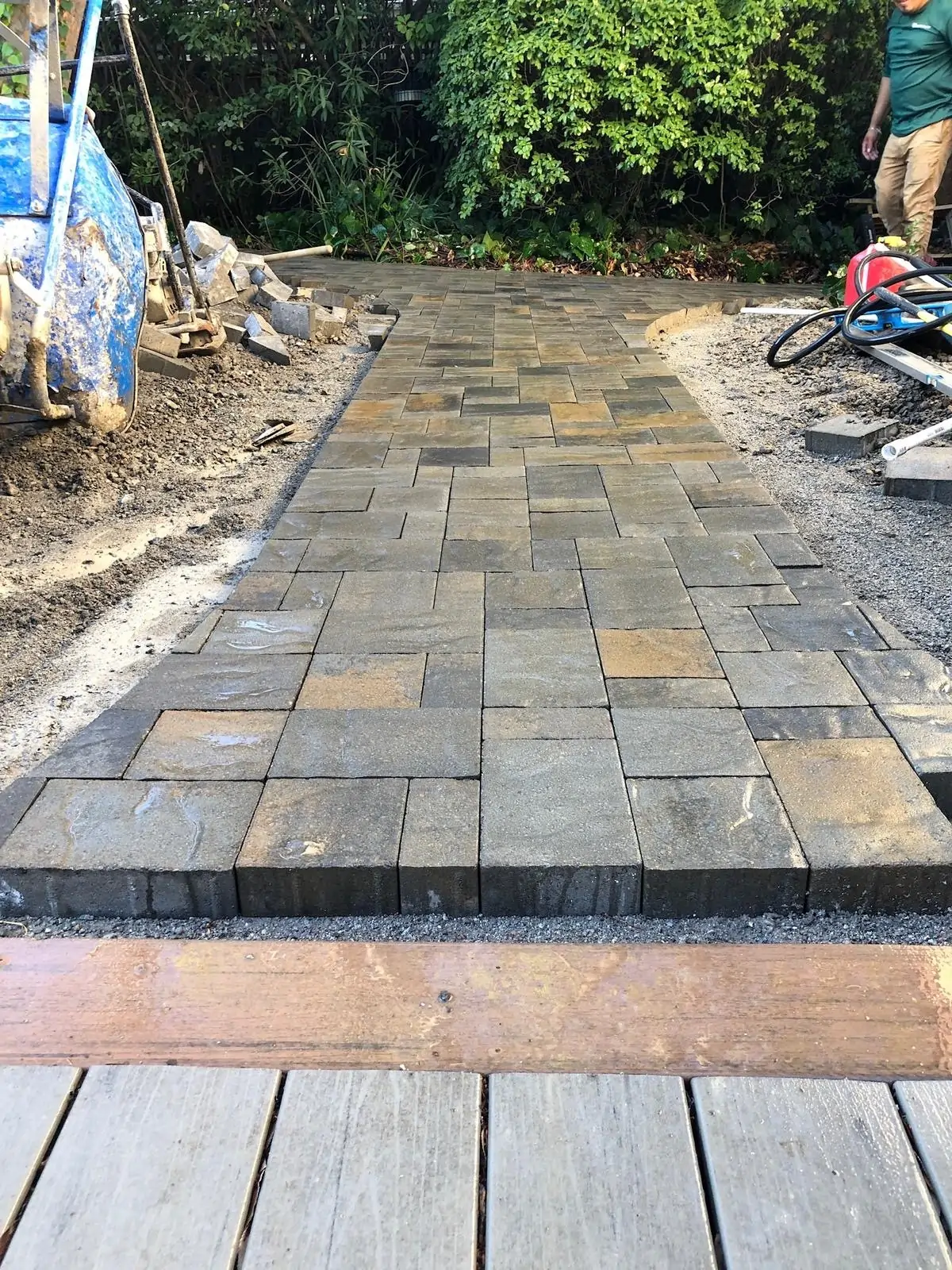 Walkway-Pavers-Service-3 1 - Three Rocks Pavers, Inc. A freshly laid path of interlocking gray pavers is bordered by construction materials, leading towards a garden area.
